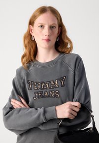 Felpa grigia con la scritta "TOMMY JEANS" ricamata in lettere a motivo leopardato; presenta un colletto a girocollo e una vestibilità rilassata. Borsa nera tenuta in mano.
