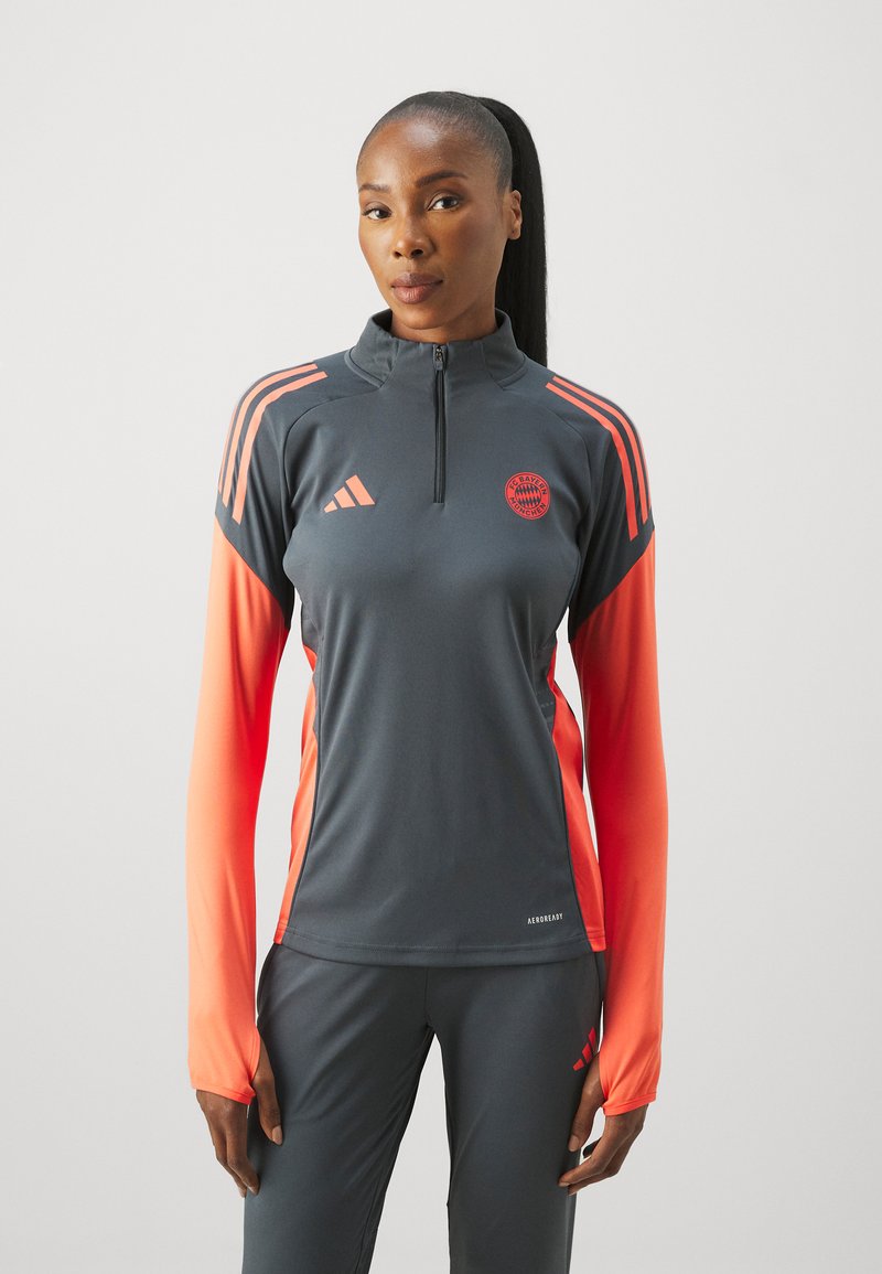 Grijze sportshirt met lange, oranje mouwen, een halve ritssluiting bij de hals en een embleem op de borst; gemaakt van een glad, rekbaar materiaal.