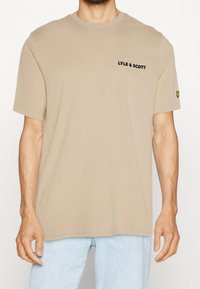 Beżowa koszulka z krótkim rękawem z napisem "LYLE & SCOTT" na klatce piersiowej i żółtym logo ptaka na lewym rękawie, noszona z jasnoniebieskimi dżinsami.