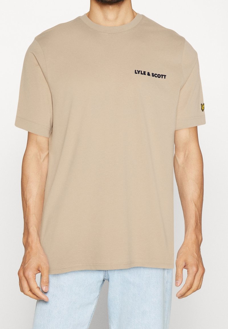 Beżowa koszulka z krótkim rękawem z napisem "LYLE & SCOTT" na klatce piersiowej i żółtym logo ptaka na lewym rękawie, noszona z jasnoniebieskimi dżinsami.