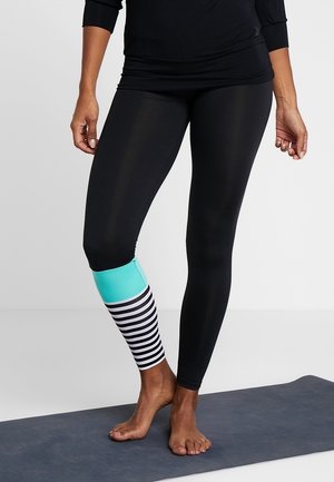 Henkilö seisoo paljain jaloin joogamatolla, päällään mustat leggingsit, joissa on turkoosi ja mustavalkoraidallinen lahkeen alaosa.