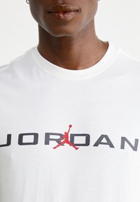 Camiseta de algodón blanca con "JORDAN" en letras negritas negras y un logo de un hombre en salto en rojo centrado debajo. Mangas cortas y escote redondo.
