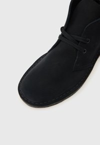 Μαύρη σουέτ μπότα τσούκα με στρογγυλεμένη μύτη, μαύρα κορδόνια και ορατή ραφή κατά μήκος της άκρης της σόλας σε άσπρο φόντο.