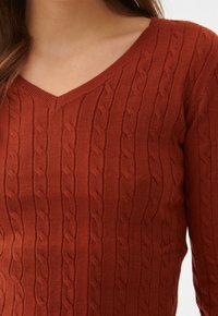 Gros plan sur une personne portant un pull en maille torsadée couleur rouille à col en V et manches longues.