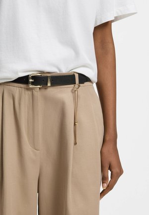 Schwarzer Ledergürtel mit goldener Schnalle und Kettendetail, getragen zu beigen Hosen und weißem Hemd, gezeigt an der rechten Körperseite.