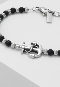 Pulsera de charm con ancla plateada que presenta cuentas negras y plateadas, cadena de metal y un cierre con una pequeña etiqueta.