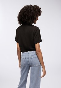 Top nero a maniche corte con una finitura testurizzata, indossato con jeans a gamba larga blu chiaro con due tasche posteriori. L'outfit è casual e morbido.