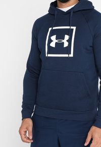Uomo che indossa un hoodie blu navy di Under Armour con un logo bianco all'interno di un quadrato sul petto, in piedi contro uno sfondo semplice.