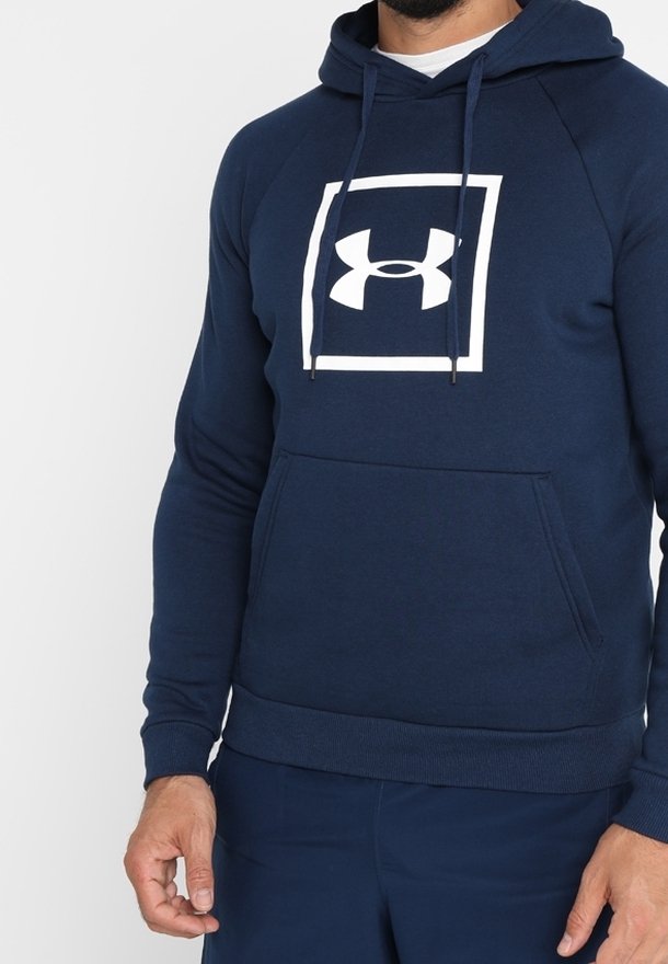 Uomo che indossa un hoodie blu navy di Under Armour con un logo bianco all'interno di un quadrato sul petto, in piedi contro uno sfondo semplice.