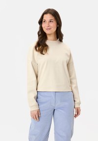 Hellbeige, verkürzter Pullover mit langen Ärmeln, Rundhalsausschnitt und gerippten Bündchen. Weiche Textur, minimalistisches Design, kombiniert mit hellblauen Hosen.