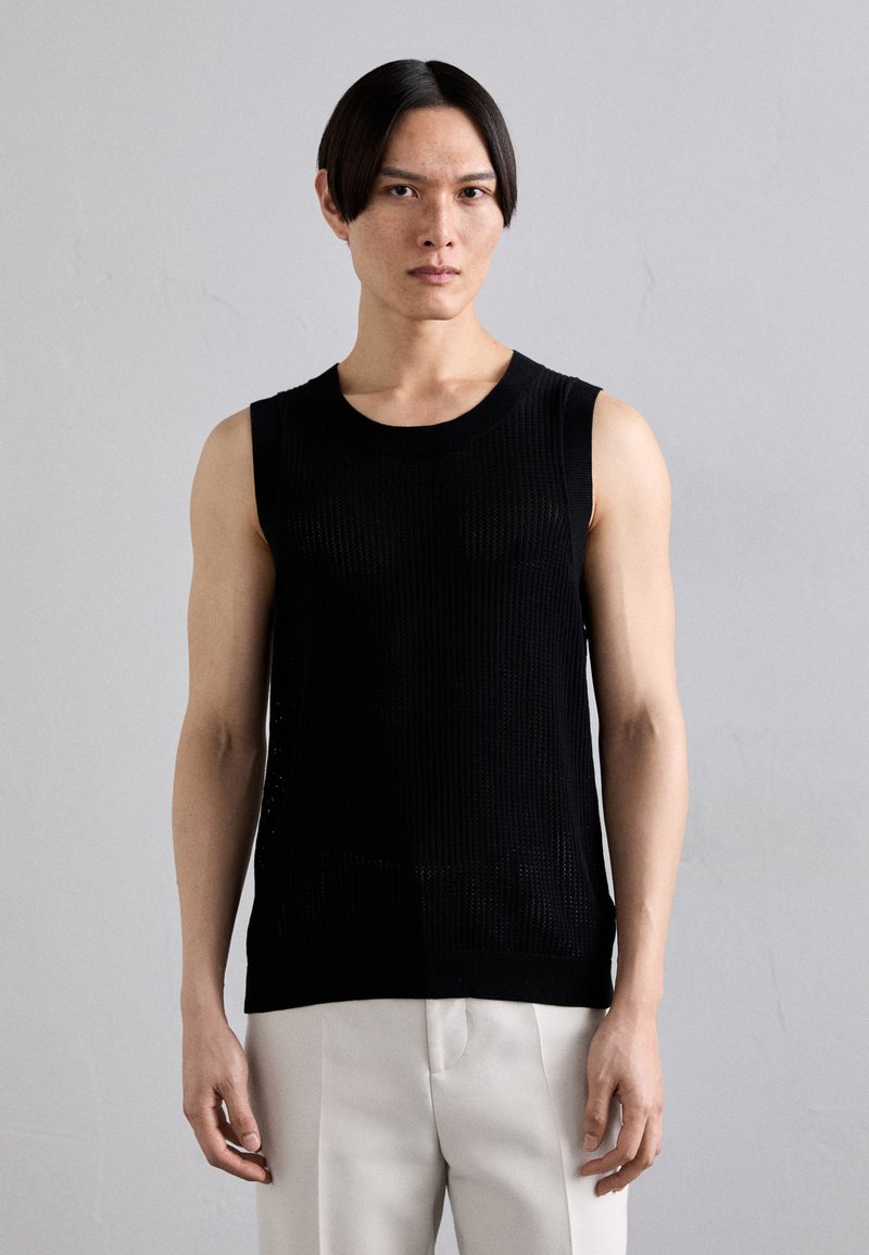 Black sleeveless ribbed knit top with a round neckline, complemented by smooth texture and a fitted silhouette. Worn with light-colored trousers.