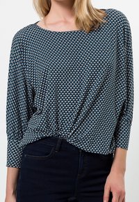 Blau gemusterte Bluse mit geometrischem Design, weichem Stoff, rundem Halsausschnitt und lockerer Passform mit hochgerollten Ärmeln, kombiniert mit dunklen Jeans.