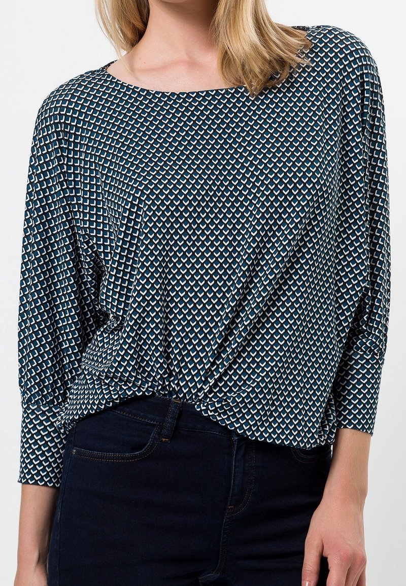 Blau gemusterte Bluse mit geometrischem Design, weichem Stoff, rundem Halsausschnitt und lockerer Passform mit hochgerollten Ärmeln, kombiniert mit dunklen Jeans.