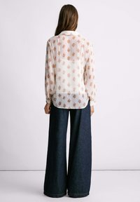 Blusa fluida color crema con motivo floreale; maniche lunghe, polsini con bottoni e orlo posteriore leggermente più lungo; abbinata a pantaloni di jeans a gamba larga.