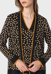 Svart tröja med beige prickar, med V-ringning och kontrasterande orange kant längs kanterna och ärmslut. Slät textur, långa ärmar.