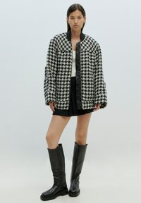Schwarze und weiße Hahnentrittmuster gepufferte Jacke mit Reißverschluss, oversized Passform, elastischen Bündchen, kombiniert mit einem schwarzen Minirock und kniehohen Stiefeln.