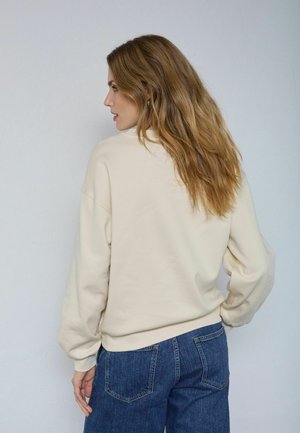 Bēšs sporta krekls ar brīvu piegriezumu, ar ribotu aproci un apakšmalu. Valkāts kopā ar zilām augstas jostasvietas džinsa biksēm. Vienkārša auduma struktūra bez redzamiem rakstiem.