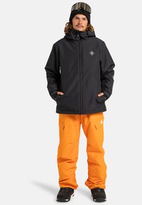 Muž v čiernej zimnej bunde, čiernej čiapke so snehovými okuliarmi, jasne oranžových snehových nohaviciach a čiernych snehových čižmách, stojí vzpriamene.