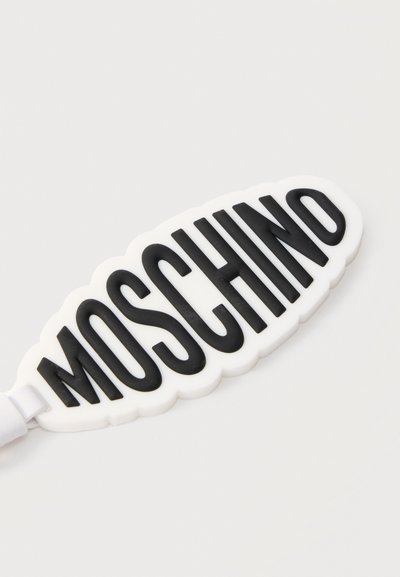 Λευκό μπρελόκ με ανάγλυφα μαύρα γράμματα που σχηματίζουν τη λέξη "MOSCHINO" σε απλό ανοιχτό φόντο.