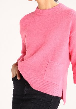 Lyserød sweater lavet af blødt stof med ribbet halsudskæring, sideslids og en lomme på forsiden. Båret med sorte jeans.
