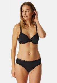 Kvinde justerer sit hår iført sort bh med underwire og matchende trusser med blonder, stående foran en ensfarvet lys baggrund.