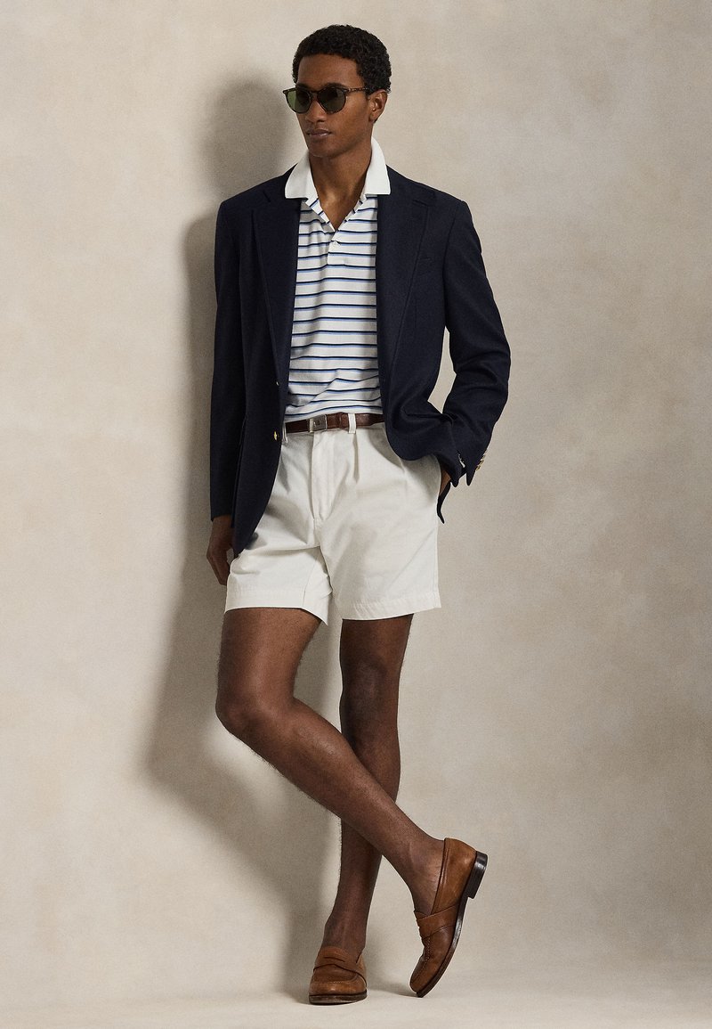 Blazer bleu marine porté sur un polo rayé, shorts beiges et mocassins marron. Accessoires : lunettes de soleil foncées. Fond texturé.