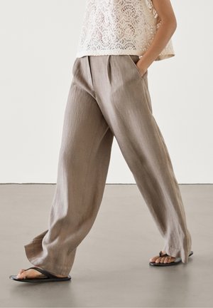 Pantalon en lin marron clair avec une coupe ample et un devant plissé. Associé à un haut en dentelle crème et des sandales noires. Design et texture minimalistes.