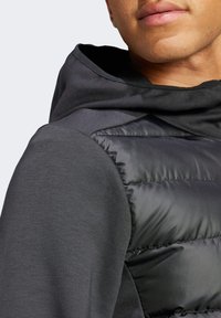 Czarna, pikowana kamizelka o gładkim wykończeniu, noszona z ciemnoszarym kapturem. Charakteryzuje się eleganckim designem i kontrastowymi teksturami na rękawach.