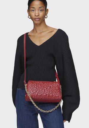 Borsa a tracolla in pelle rossa trapuntata con tracolla a catena, caratterizzata da una texture liscia e un motivo arrotondato e astratto. Abbinata a un maglione nero a coste.