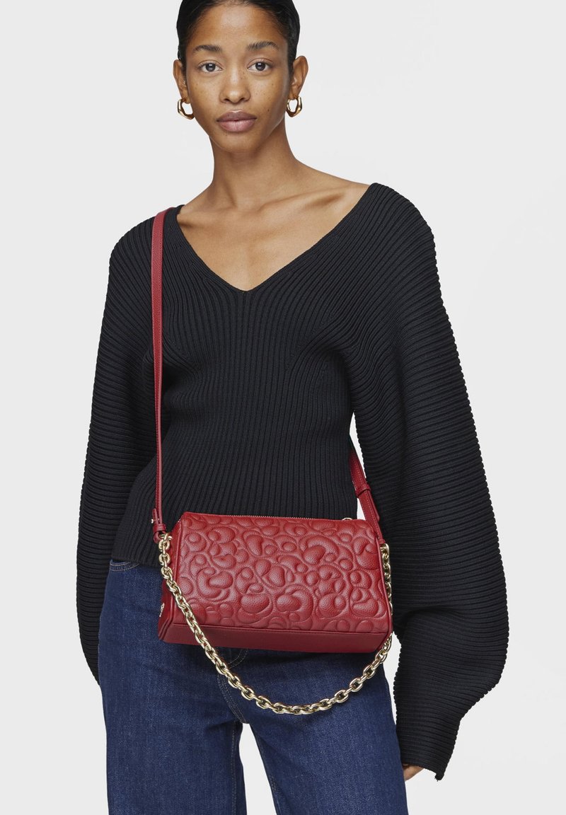Czerwona pikowana skórzana torebka na ramię z łańcuszkiem, o gładkiej fakturze i zaokrąglonym, abstrakcyjnym wzorze. Stylizowana z czarnym prążkowanym swetrem.