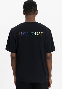 Zwarte katoenen t-shirt met meerkleurige geborduurde tekst op de achterkant die "LEEF VANDAAG" zegt. Korte mouwen met een ruime pasvorm.