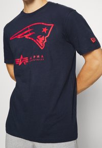 Námořnická modrá tričko vyrobené z bavlny s červenou grafikou stylizované hlavy a hvězdy, s nápisem "ALPHA INDUSTRIES" a akcenty na rukávech.