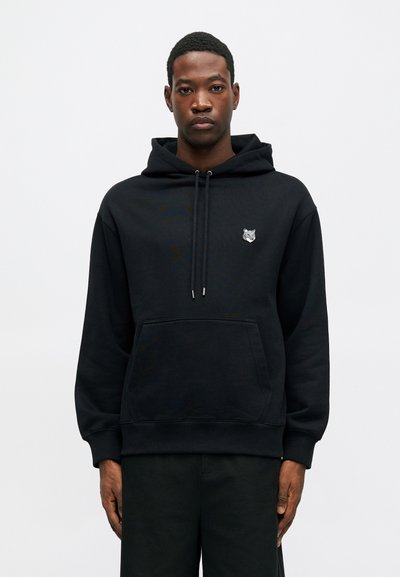 Hoodie en coton noir avec une poche avant, un capuchon à cordon de serrage et un petit design de loup brodé sur la poitrine. Texture lisse, coupe décontractée.