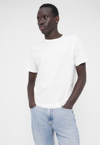 White cotton t-shirt with a crew neckline, paired with light blue denim jeans. Model stands with hands in pockets, on a plain white background.