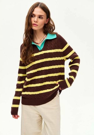 Młoda kobieta z długimi brązowymi włosami, ubrana w brązowo-żółty sweter w paski z zielonym kołnierzem, beżowe spodnie i koralikowy naszyjnik.