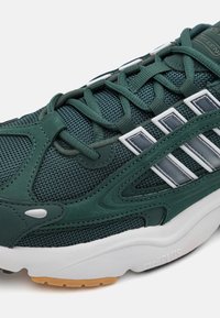 Green mesh and suede sneaker featuring white and silver accents, textured upper, padded tongue, and a thick white and gum rubber sole.