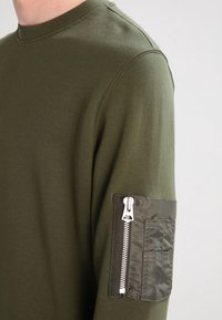 Maglietta a maniche lunghe color verde oliva con scollo rotondo realizzata in un tessuto testurizzato, dotata di una tasca con zip sulla manica sinistra in un tessuto contrastante.