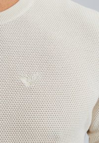 Lichtbeige gebreide trui met een textuurpatroon, geribbelde ronde hals en een klein geborduurd embleem op de borst.