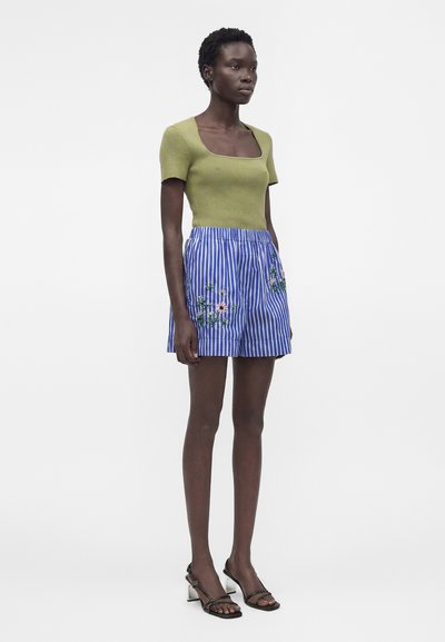 Femme debout portant un haut vert ajusté à manches courtes, un short rayé bleu et blanc avec broderie florale et des sandales noires à talons.