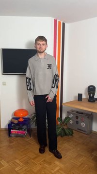 Helehall hall kaunistatud mustade detailide ja logoga kootud kampsun, millele on sobitatud mustad lahedalt lõigatud püksid ja tumepruunid kingad. Siseruumides.