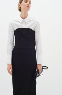 Rochie neagră structurată, cu un overlay dintr-o cămașă albă cu mâneci lungi, având un corset ajustat și o fustă dreaptă. Accesoriu: posetă mică neagră.