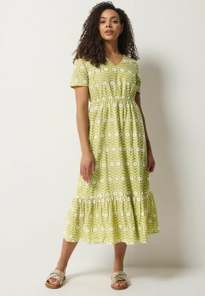 Femme aux cheveux bouclés portant une robe midi vert clair à motifs floraux blancs et ourlet à volants, associée à des sandales beiges tressées.