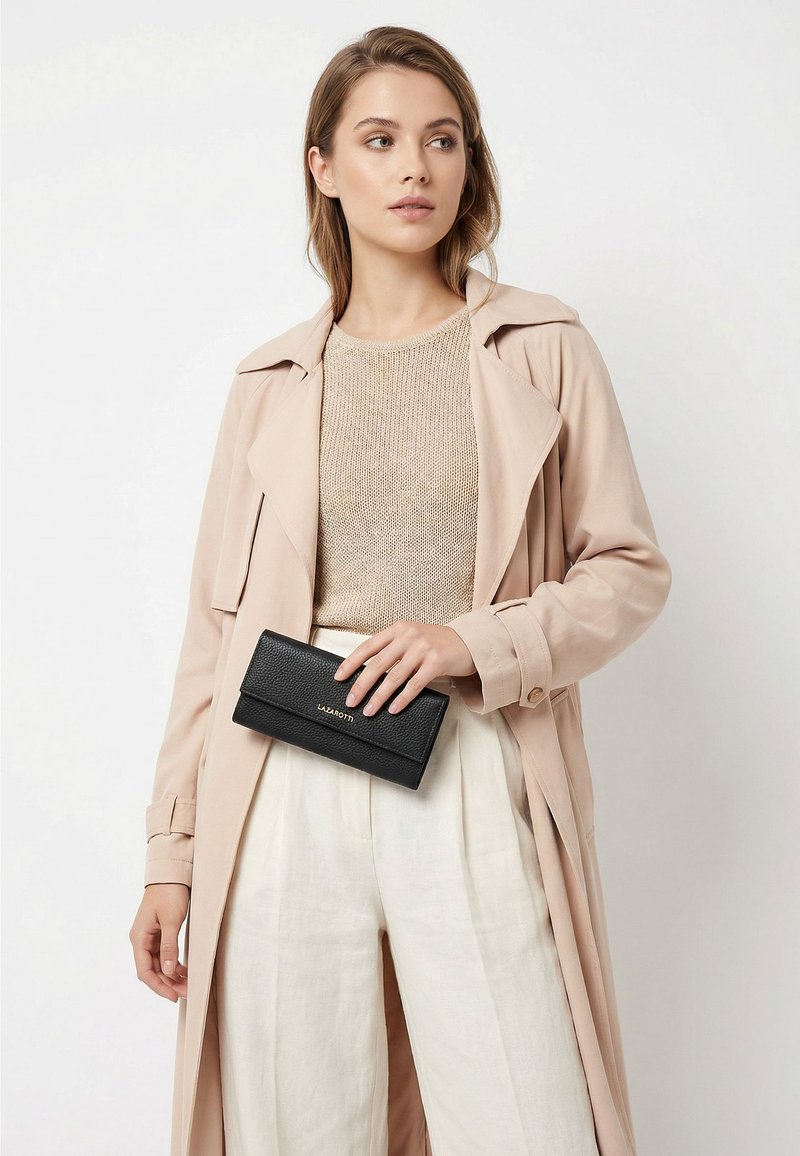 Mujer con abrigo beige y pantalones claros sosteniendo una cartera negra texturizada con letras doradas frente a un fondo blanco.