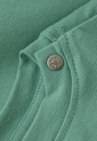 Tissu vert avec des coutures détaillées et un bouton en argent présentant un trèfle à quatre feuilles embossé.