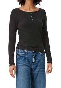 Frau trägt ein schwarzes, langärmeliges, geripptes Shirt mit drei Knöpfen am Ausschnitt und blaue Jeans, steht vor einem weißen Hintergrund.