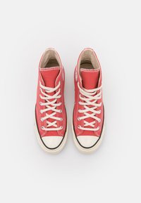 Červené vysoké tenisky vyrobené z plátna s bílými tkaničkami, gumovou špičkou a černou gumovou podešví. Obsahuje logo na vnější straně.