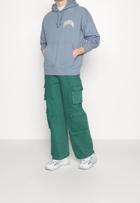Světle modrá mikina s kapucí, přední kapsou a logem, spárovaná se zelenými cargo kalhotami s několika bočními kapsami. Bílé tenisky dokončují vzhled.