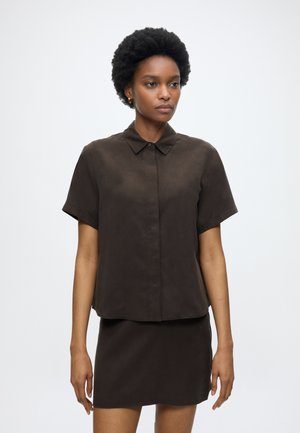 Femme aux cheveux courts et bouclés portant un chemisier à manches courtes marron foncé avec col et une jupe assortie, debout devant un fond clair uni.