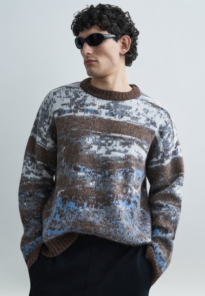 Gestrickter Pullover in Braun-, Grau- und Blautönen. Verfügt über einen Rundhalsausschnitt, lange Ärmel und ein strukturiertes Muster. Wird über dunkler Hose getragen.