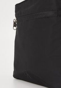 Sac en nylon noir avec fermeture zippée sur le dessus, doté d'un logo embossé discret et d'une tirette en argent, mettant en avant un design élégant et fonctionnel.